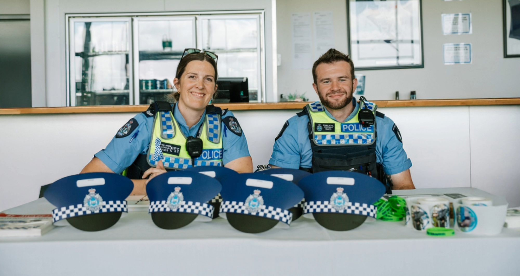 Two police officers in uniform sitting behind a desk at the 2025 Mandurah Community Safety Expo with information pamphlets and cardboard police hats 
